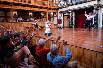 The audience plays Ophelia's Super Ego with hands waving in the air  directed by Ginna Hoben, kneelg at the front of the stage wearng a white smock and blue jeans with a red carnation pinned to her left lapel, and Jenny Bennet standing at he back of the stage wearing white shirt, black and white Elizabehtn knee britches, and suspenders.