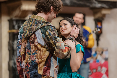 Romeo with back to us is wearing a multicolor hoodie, his right hand stroking her hair as she gaes up into his eyes, her left hand on his right arm. She's wearing a turquoise dress. A man in a blue and yellow letter jacket is playing guitar at the back of the stage.