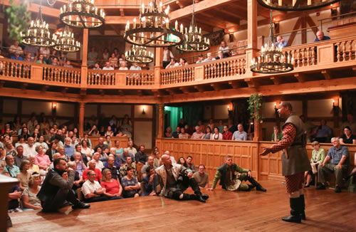 Berowne standing on the stage is in breen short coat over red and gold patterend Elizabetan jacket and knee britches with white stockings and black boots. His hands are outstretched. Sitting at the front of the stage, facing him and backs to the audience, each with an arm on a knee, are Dumaine, Ferdinand, and Longaville. All around (including sitting on stools on the stage) are the audience filling the all-wood, two-tier  theater with more than a dozen iron chandeliers hanging above and flower vines on the posts.