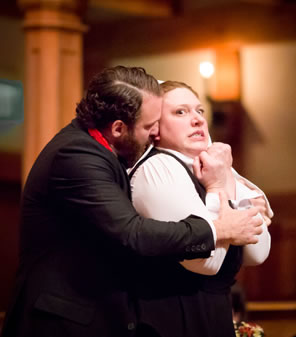 Angelo in black coat and medallion on red ribbon around his neck clasps Isaballa from behind, she with her arms bent tight, fists close to her chin, a look of fear on her face