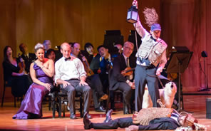 Hippolyta in purple gown and crown sits next to Theseus in white suit shirt and bowtie in front of members of the orchestra string section, all in black formal wear, leaning over to see, bottom in whicker breastplate and funny helmet lies on the floor as Moonshine in flack-jacket, baseball cap with thorn bush, holding up a lantern and leash on white dog stands over him.