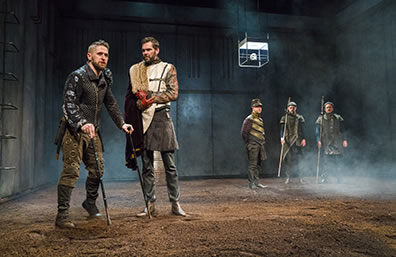 Production photo of Richard, wearing a black jacket and brown pants with boots, leaning on his two cans, talking with Clarence, in a white jerkin, skirt, leather pants, and a cape on his right shoulder. Three men stand guard in the background. 