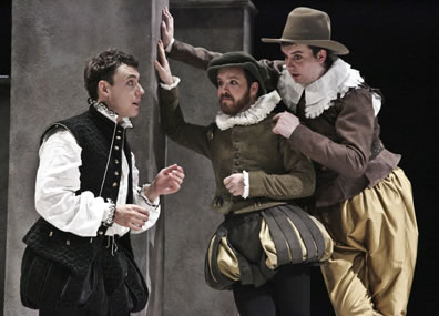 Hamlet in bulging black Elizabethan-style breeches, black vest and high collar white shirt to the left talks with Rosencrantz in green and gold breeches and jacket with white ruff collar dog cap and Guildenstern in gold pants brown jacket and lace collar with country brimmed hat, both leaning against a wall