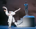 Hermione in white gown, on point with her right leg, left leg extended back and arms arching over her head, Leontes in white overcoat, arms wrapped around her waist and under her extended leg, head leaning into the crook of her back, a statue of Mamillius on a white pedestal in the background