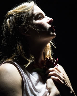 Kimberly Gilbert clutching a flower to her breast looks up to the right, deep shadow on her face