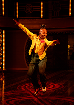 SIG-Play_On-Jester Jester with orange shirt and jacket, carnation in jacket button hole, dark pants, and black and white tap shoes is airborne with outstretched arms as he dances. Photo by Daniel Rader