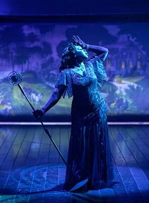 SIG-Play_On-Lady_Liv Lady Liv in blue fancy dress and thin cape around shoulders, leans back dramatically as she stands in a blue spotlight  with gloved left hand to her forehead and her outstreched right hand holding the stand of an old microphone. Curier and Ives lithograph of "A Home on the Mississippi" is in the background. Photo by Daniel Rader