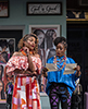 STC-Madams_Page_Ford-015 Madam Page wearing a bare-shoulder orange patterned blouse and orange and red  and white geometric patterned dress looks over at the two letters Madam ford, in baggy blue blouse, a dozen or more colorful bead necklaces hanging about her neck, and tight purple and black patterend yoga pants. Behind are signs of braided hair on the wall. and at the top a sign that says "God is Good all the time"