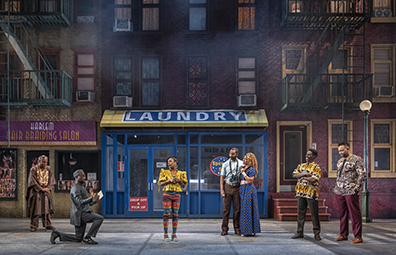 STC-Merry_Wives_Street-091 Characters in various West African-styled clothing (except the Pages, he in  common American short-sleeve shirt and pants with binoculars hanging around his neck from birdwatching, and she in floor length blue dress with small yellow X's) watch Ford's contritio in front of a street scene of the salon and laundryomat with tenements on either sice and a street lamp to one side.