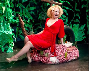 Olvia in revealing red dress sitting on coral reef amid seaweed.